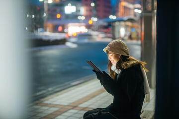 Fototapeta premium Asian woman remote working freelance on digital tablet while waiting for bus in winter night. Woman traveler enjoy urban lifestyle travel in the city with using wireless technology and gadget device.