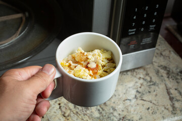 A view of a hand holding a mug of instant breakfast bowl next to a microwave.