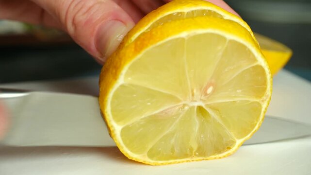 Close up hand holding, slicing fresh ripe yellow lemon with sharp kitchen knife on white cutting board, preparing healthy ingredient for cooking, refreshing drink. Slicing fresh juicy lemon with knife