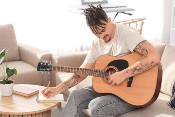 Young man with dreadlocks and guitar writing in notebook on sofa at home