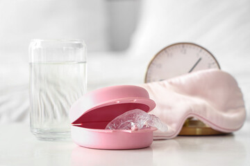 Box with occlusal splint, glass of water and sleeping mask on table in bedroom, closeup