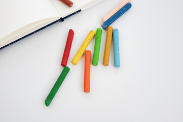 Colorful chalk sticks on white surface next to open notebook. 