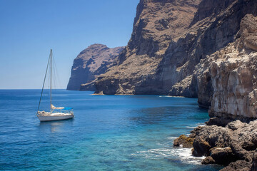 Fototapeta premium Sailboat floating on clear blue sea near rocky cliffs