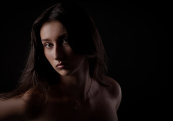 Fototapeta premium Studio portrait of young woman with long brown hair and bare shoulders, highlighted by dramatic lighting against a black background