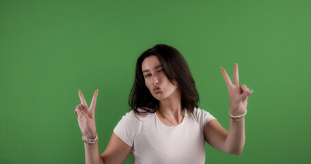 Young woman making peace signs with both hands and kissing on a green screen background