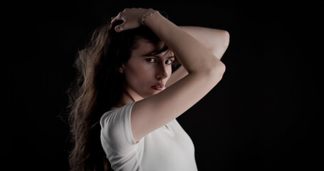 Studio portrait of a young woman with long brown hair wearing a white t shirt, posing with her hands on her head