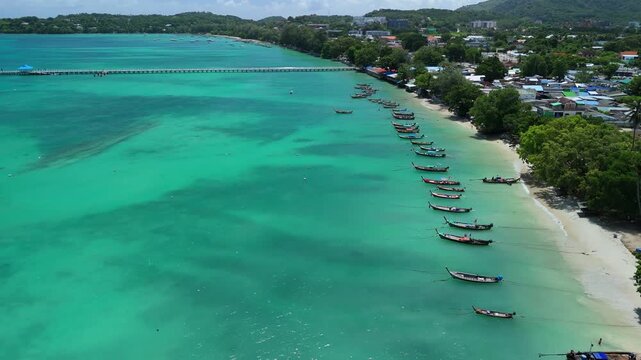 Drone pushes back above moored longtail boats along turquoise Rawai Beach, Phuket