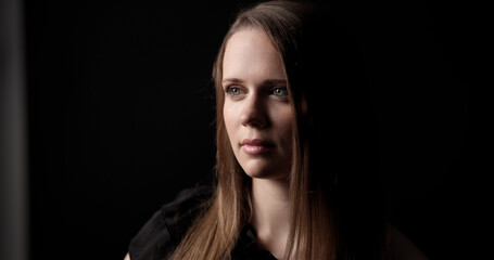 Naklejka premium Close up portrait of a young woman with long brown hair, wearing a dark shirt, looking away from the camera with a thoughtful expression in a studio setting with dark background