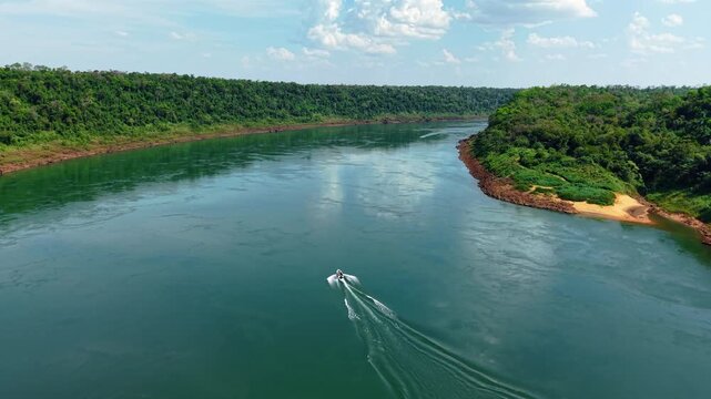 Boat gliding along the Paran&aacute; River near the border with Paragua, framed by forest canopy and Misiones&rsquo; red soil&mdash;captured by descending drone, scene blends motion, ecology, and tropical tranquility.