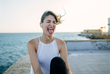 Woman listening music with earphones and sticking out tongue by the sea