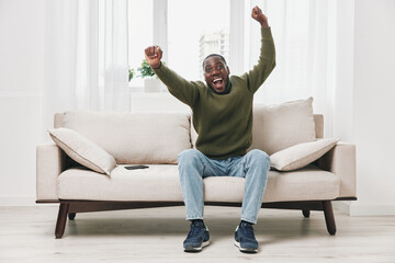 Happy African American man celebrating success while sitting on a cozy sofa, wearing a casual green...