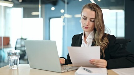 Corporate professional businesswoman in a black suit and white shirt, focused while working on laptop and reviewing documents in modern office environment. Demonstrates diligence corporate activity. - Powered by Adobe