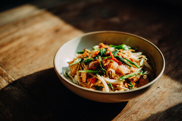 Authentic Thai Pad Thai noodles served in a ceramic bowl on rustic wooden table, garnished with bean sprouts, chives, and lime. Traditional Asian street food dish in warm natural lighting.