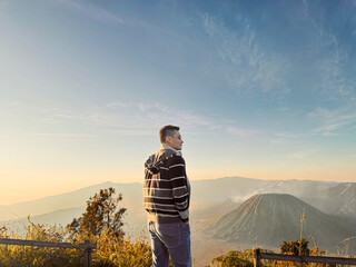 A guy posing with Mount Bromo and Mount Batok at the background, with beautiful and colorful sunrise sky in the early morning. In Bromo Tengger Semeru National Park, Indonesia. Travel photography