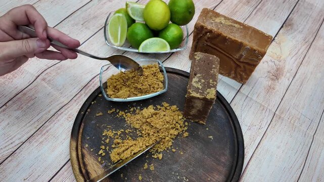Close-up shot hand scooping and measuring grated panela (papel&oacute;n) with a spoon, ingredients: raw sugar blocks and the fresh green limes, ready for traditional papel&oacute;n con lim&oacute;n beverage
