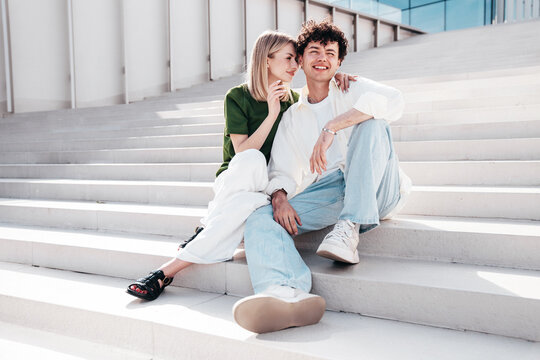 Smiling beautiful woman and her handsome boyfriend. Couple in casual summer clothes. Happy cheerful family. Female and man having fun. They posing in the street in sunny day. Sit at stairs - Powered by Adobe
