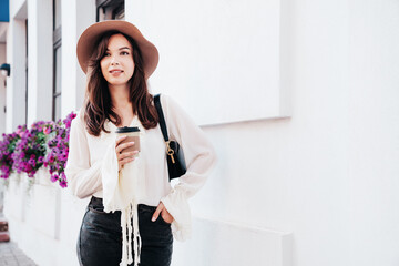 Lifestyle portrait of young stylish woman dressed in white blouse. Model walking with coffee cup in old town in sunny day. Beautiful smiling female in hat. Cheerful and happy. Takeaway coffee
