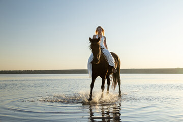 A woman in a flowing white dress rides her horse through the shimmering waters of Kuyalnik Lake, bathed in the golden evening light.
