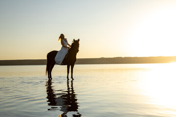 A woman in a white dress rides her horse through Kuyalnik Lake at sunset, glowing in the golden light with serene water reflections.