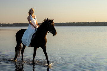 A woman in a flowing white dress rides her horse through the calm waters of Kuyalnik Lake at sunset, embodying elegance and freedom.