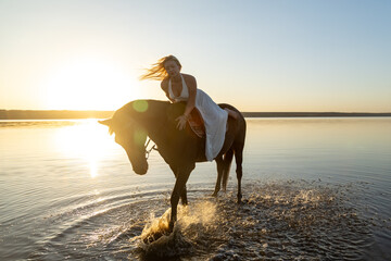 A woman in a flowing white dress leans close to her horse, water splashing under its hooves at Kuyalnik Lake during sunset.