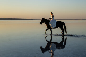 A woman in a flowing white dress rides her horse across Kuyalnik Lake, their reflection glowing in...