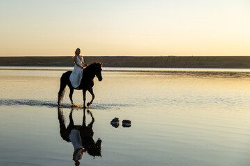 A woman in a white dress rides her horse along Kuyalnik Lake, their reflection mirrored perfectly in the calm sunset waters.