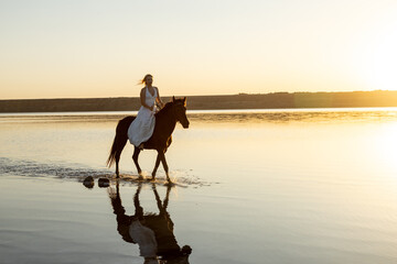 A woman in a white dress rides her horse across Kuyalnik Lake, bathed in the warm glow of the setting sun with their reflection shimmering in the water.