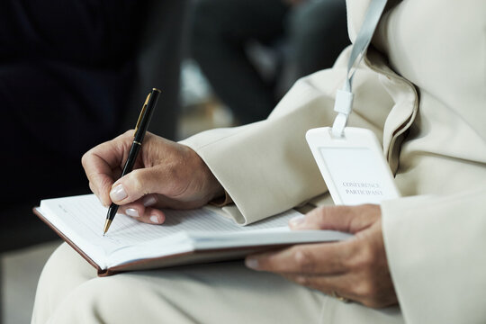 Middle aged unrecognizable woman writing in notebook while attending conference, holding pen in right hand and wearing visible name badge, focusing on taking notes during event
