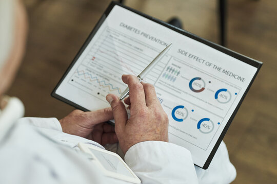 Senior male doctor holding pen and reviewing medical chart with diabetes prevention and medicine side effect statistics, sitting indoors with partial coat visible - Powered by Adobe