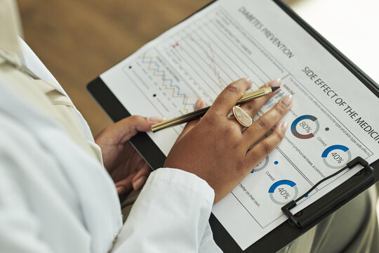Closeup of Black female medical professional holding clipboard, analyzing diabetes prevention chart and side effect statistics, wearing coat, sitting at desk, hand visible