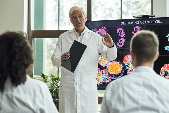 Senior Caucasian male doctor explaining cancer cell destruction process to diverse group of young colleagues in conference meeting, holding folder while standing in front of scientific display screen