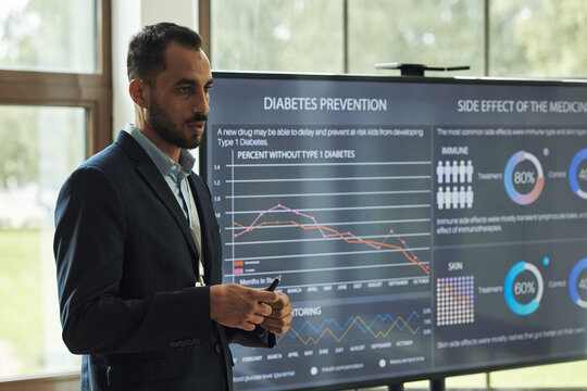 Young polyethnic man presenting diabetes prevention data on large digital screen, holding remote control, standing in front of medical statistics and charts during seminar in hospital - Powered by Adobe