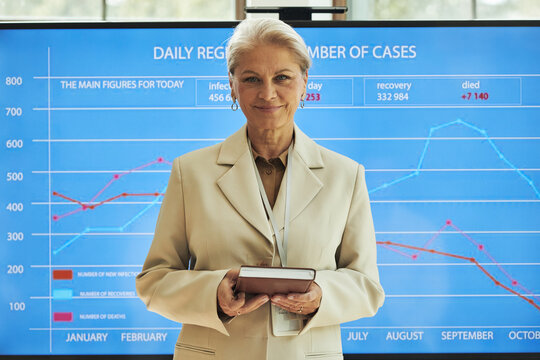 Portrait of senior Caucasian woman standing in front of large digital chart holding notebook, smiling confidently, presenting statistical data on daily registered number of medical cases at conference