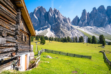 wooden chalet and green meadow with fence under mountain Odle group in Dolomites in Italy