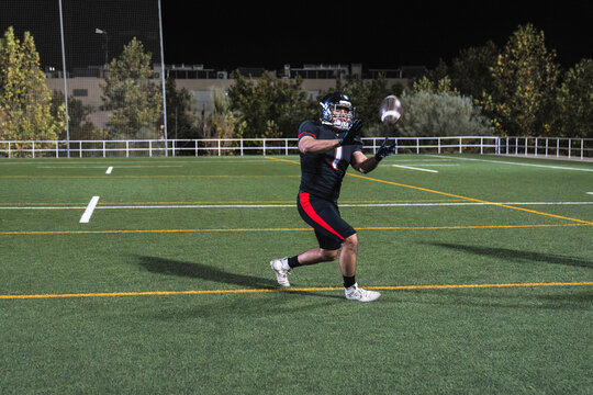 Male athlete in uniform playing american football, actively catching the ball during night game on green field