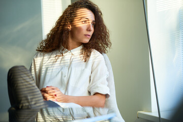 Attractive long-haired curly woman in white shirt sitting in soft chair in her office, looking through window thoughtfully with her hands clasped on belly