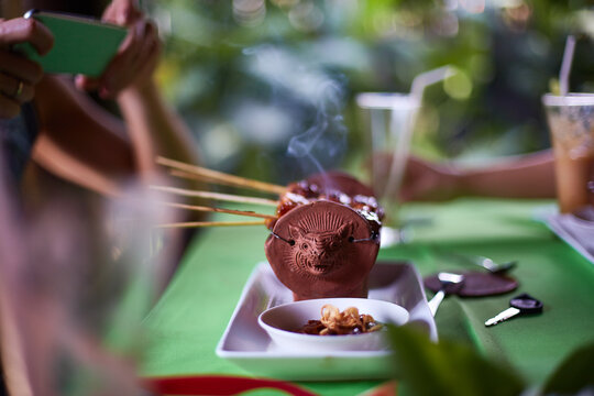 Hands of young adults holding smartphones and photographing traditional grilled skewers served in decorative clay pot with smoke rising, food and drinks on green table, outdoor setting