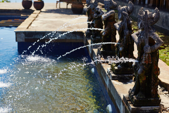 Row of traditional stone statues shaped as mythological figures spouting streams of water into outdoor pool, statues aligned along pool edge with clear water splashing