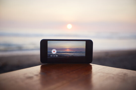 Smartphone recording sunset over ocean, device placed on wooden surface with camera capturing scenic view, horizon and sun reflecting on water, tranquil outdoor setting