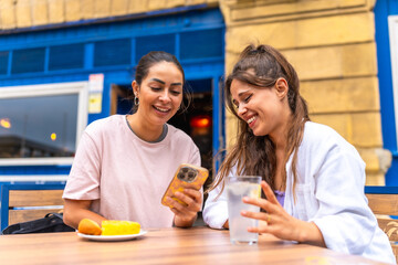 Two happy women sharing funny content on a smartphone while having breakfast at a bar