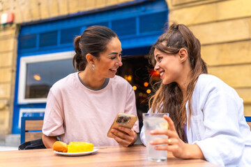 Two smiling women sharing a moment at outdoor cafe, browsing smartphone