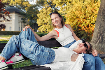 Pair smiling happy young adult women enjoy sunny day together sit relaxing on bench in city park outdoors. Female persons wearing casual denim white tops portrait. Best friends friendship outside