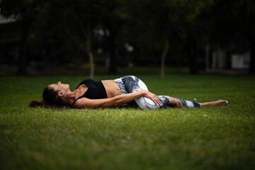 Woman performing supta matsyendrasana spinal twist yoga pose in park