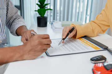 A businesswoman in a yellow suit is consulting with a male client about car purchase contracts, showing documents and car models on the desk to finalize agreement and transaction.