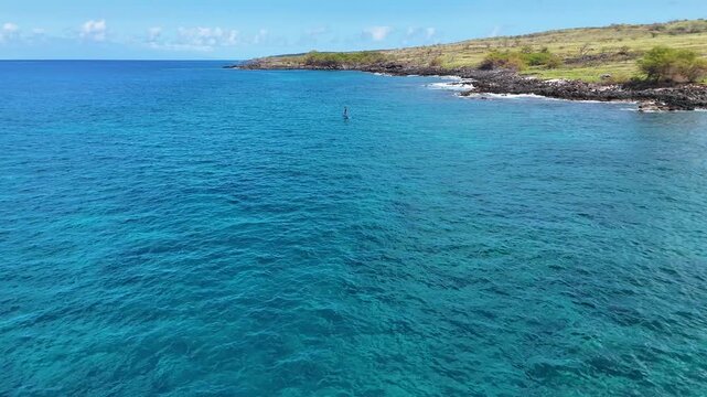 Aerial view of person paddleboarding on crystal-clear turquoise water near the rocky coast, displaying a striking contrast of colors, Hawi, Hawaii, United States.