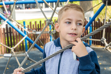 Cute boy with a modern haircut playing and climbing on a rope net structure at an outdoor playground, looking away, close-up