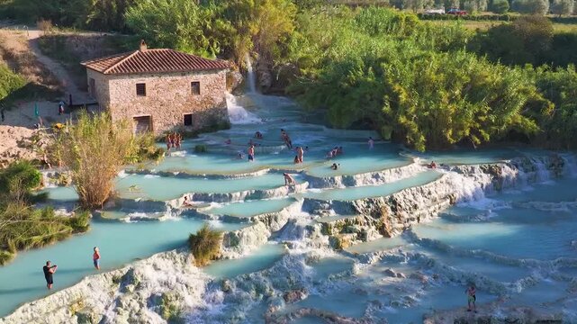 Aerial view of the Saturnia hot springs, where turquoise waters cascade over tiered rocks, contrasting with the lush green vegetation, Saturnia, Toscana, Italy.