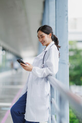 Smiling doctor using smartphone leaning on railing in hospital corridor