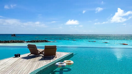 The infinity pool meets the turquoise ocean under a bright blue sky with two lounge chairs on the wooden deck.